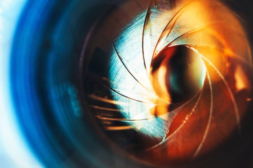Description : Gros plan artistique d'un diaphragme d'objectif de vidéoprojecteur ou de caméra, montrant les lamelles métalliques ouvertes et des reflets lumineux bleus et orangés, illustrant la précision optique et la résolution.
