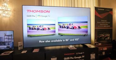 Grand téléviseur Thomson QLED Pro Google TV exposé lors d'un salon professionnel avec mention Now also available in 85 et 100 pouces, entouré de packagings produit et d'un kakémono promotionnel