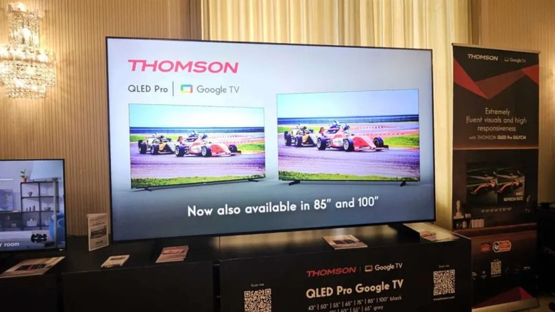 Grand téléviseur Thomson QLED Pro Google TV exposé lors d'un salon professionnel avec mention Now also available in 85 et 100 pouces, entouré de packagings produit et d'un kakémono promotionnel