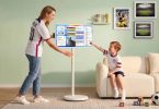 Apolosign Smart Portable TV Gen 2 32 pouces 4K sur pied à roulettes, mère en maillot équipe USA foot et enfant interagissant avec l'interface dashboard tactile dans salon contemporain mur vert, photos foot encadrées Coupe du Monde 2026 et ballon de football
