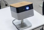 Projecteur Epson Lifestudio Flex en finition plateau supérieur bois clair veiné, façade noire avec objectif principal allumé en bleu lumineux et bandeau enceinte BOSE en tissu gris, posé sur stand pivotant aluminium