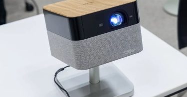 Projecteur Epson Lifestudio Flex en finition plateau supérieur bois clair veiné, façade noire avec objectif principal allumé en bleu lumineux et bandeau enceinte BOSE en tissu gris, posé sur stand pivotant aluminium