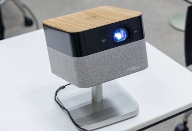 Projecteur Epson Lifestudio Flex en finition plateau supérieur bois clair veiné, façade noire avec objectif principal allumé en bleu lumineux et bandeau enceinte BOSE en tissu gris, posé sur stand pivotant aluminium