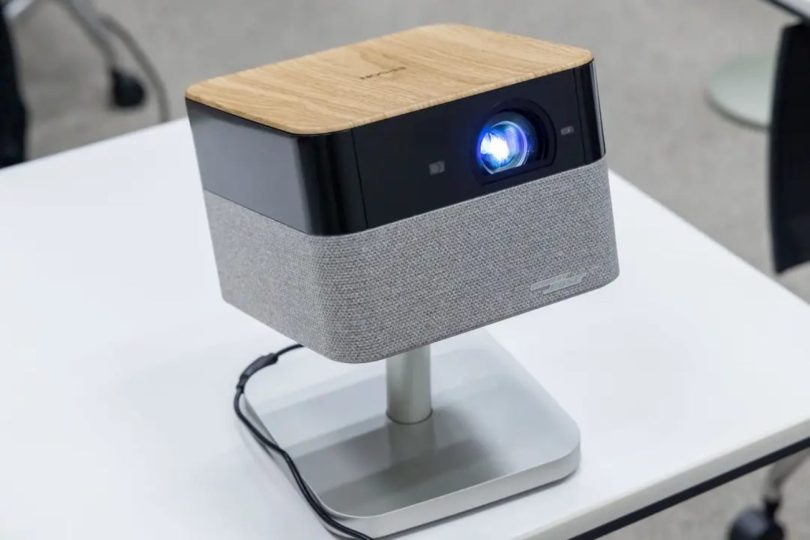 Projecteur Epson Lifestudio Flex en finition plateau supérieur bois clair veiné, façade noire avec objectif principal allumé en bleu lumineux et bandeau enceinte BOSE en tissu gris, posé sur stand pivotant aluminium