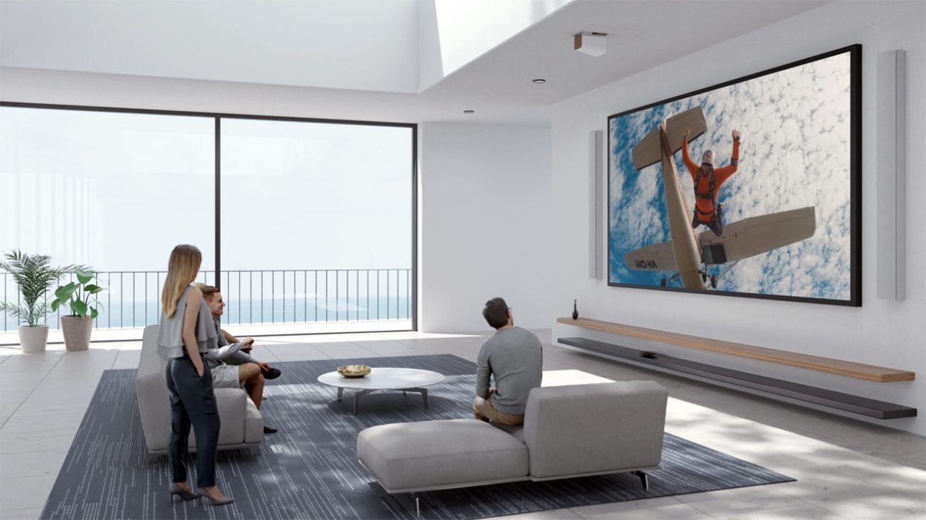 Mise en situation d'un home cinéma haut de gamme avec projecteur laser Epson installé au plafond, projetant une image très grand format (saut en parachute en plein ciel) sur mur blanc, trois spectateurs en discussion dans salon contemporain ouvert sur vue mer
