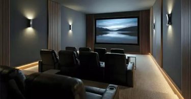 Salle de cinéma maison haut de gamme custom installation avec grand écran de projection rectangulaire montrant un paysage de lac brumeux au crépuscule, deux rangées de fauteuils club en cuir marron foncé inclinables avec porte-gobelets disposés face à l'écran, panneaux acoustiques muraux en bois lamellé vertical sur fond gris ardoise, appliques murales LED design éclairage tamisé, sol moquette beige et bandeau LED encastré au sol, ambiance cinéma privé luxueux conçu pour enceintes Fyne Audio Home Theatre Monitor IsoFlare placées derrière l'écran acoustiquement transparent