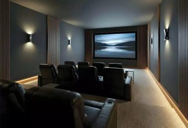 Salle de cinéma maison haut de gamme custom installation avec grand écran de projection rectangulaire montrant un paysage de lac brumeux au crépuscule, deux rangées de fauteuils club en cuir marron foncé inclinables avec porte-gobelets disposés face à l'écran, panneaux acoustiques muraux en bois lamellé vertical sur fond gris ardoise, appliques murales LED design éclairage tamisé, sol moquette beige et bandeau LED encastré au sol, ambiance cinéma privé luxueux conçu pour enceintes Fyne Audio Home Theatre Monitor IsoFlare placées derrière l'écran acoustiquement transparent