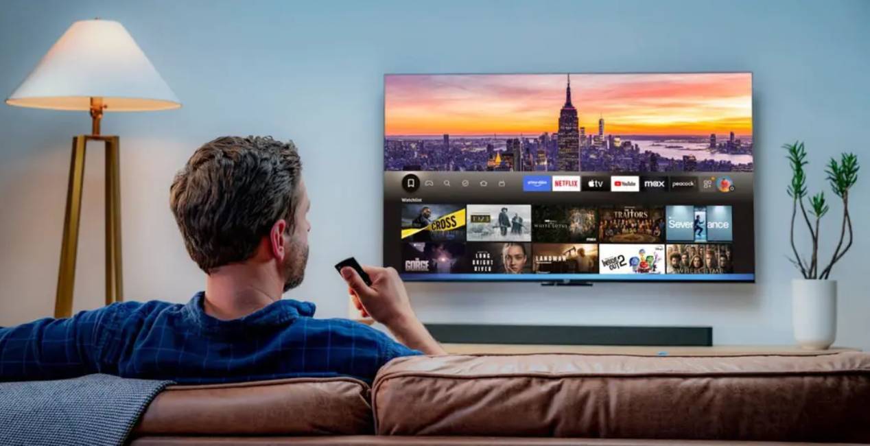 Homme installé sur un canapé dans un salon, télécommande en main, face à un grand écran affichant la page d'accueil de l'interface Fire TV avec les applications de streaming Prime Video, Netflix, Apple TV, YouTube, Max et Peacock