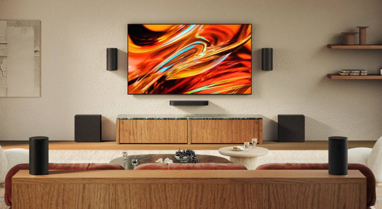 Système Sony BRAVIA Theatre Trio en configuration complète 5.2.4 dans un salon contemporain : enceinte centrale sous l'écran mural, deux satellites frontaux fixés au mur, deux caissons de basses SA-SW8 de part et d'autre du meuble bois et deux enceintes arrière cylindriques Rear 9 encadrant le canapé.
