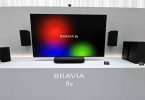 Système audio sans fil Sony BRAVIA Theatre Trio présenté sous un très grand écran : enceinte centrale compacte posée sur meuble, deux enceintes cylindriques gauche et droite encadrant l'image, caisson de basses et enceintes arrière du pack home cinéma 360 Spatial Sound Mapping.
