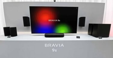Système audio sans fil Sony BRAVIA Theatre Trio présenté sous un très grand écran : enceinte centrale compacte posée sur meuble, deux enceintes cylindriques gauche et droite encadrant l'image, caisson de basses et enceintes arrière du pack home cinéma 360 Spatial Sound Mapping.