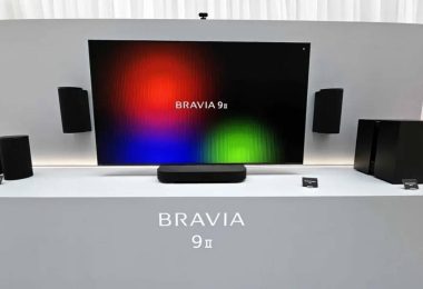 Système audio sans fil Sony BRAVIA Theatre Trio présenté sous un très grand écran : enceinte centrale compacte posée sur meuble, deux enceintes cylindriques gauche et droite encadrant l'image, caisson de basses et enceintes arrière du pack home cinéma 360 Spatial Sound Mapping.