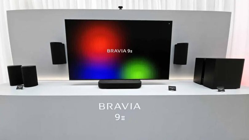 Système audio sans fil Sony BRAVIA Theatre Trio présenté sous un très grand écran : enceinte centrale compacte posée sur meuble, deux enceintes cylindriques gauche et droite encadrant l'image, caisson de basses et enceintes arrière du pack home cinéma 360 Spatial Sound Mapping.