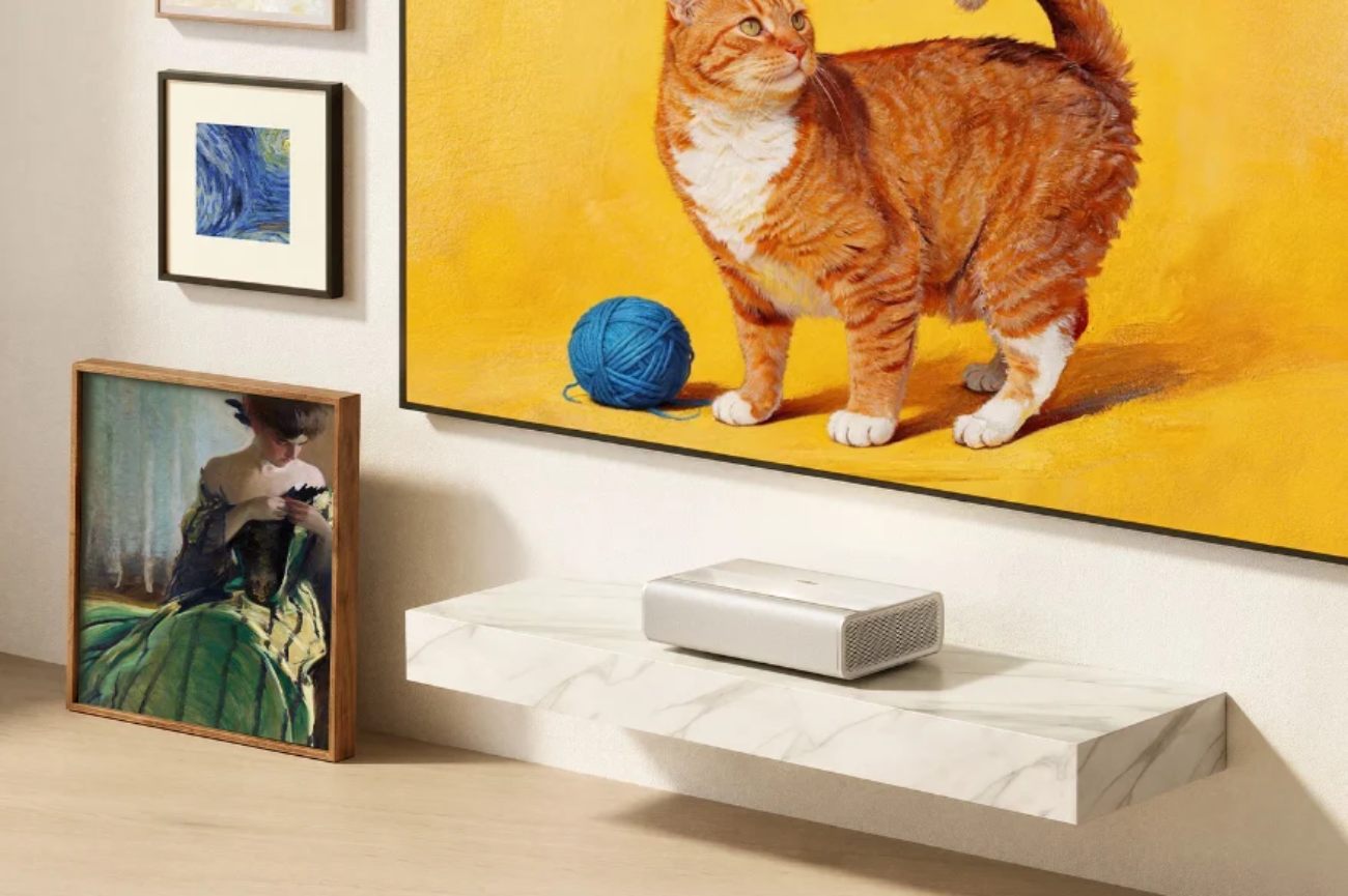 Mise en scène lifestyle du XGIMI Mira 4K Pro projetant une œuvre d'art numérique représentant un chat tigré roux orange avec pelote de laine bleue sur fond jaune éclatant en mode Light Shadow Gallery, projecteur ultra-courte focale en finition argentée posé sur étagère murale flottante en marbre blanc, intérieur galerie d'art domestique avec cadres physiques classiques disposés autour Van Gogh et portrait XIXe, sol en parquet bois clair scandinave