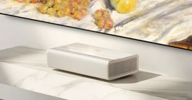XGIMI Mira 4K Pro projecteur ultra-courte focale en finition argentée minimaliste posé sur meuble TV en bois noyer foncé contre un mur projetant une œuvre d'art numérique aux teintes verts dorés en mode Light Shadow Gallery, faisceau lumineux orange visible au sommet de l'appareil, esthétique salon haut de gamme contemporain
