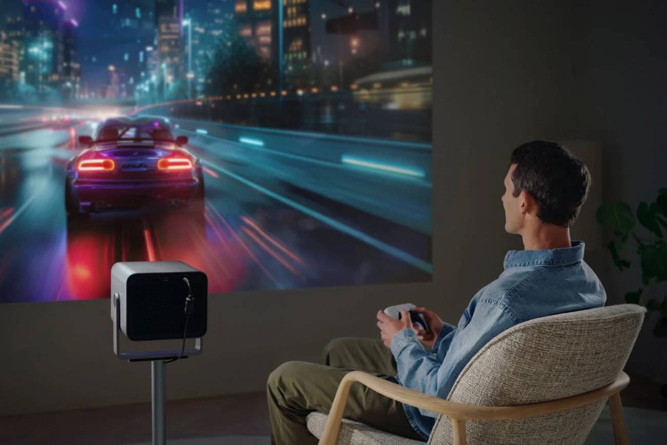 Joueur installé dans un fauteuil, manette en main, devant une grande image projetée par le Leica Cine Compact 1 posé sur un trépied dans un salon sombre, affichant un jeu de course automobile nocturne aux néons colorés