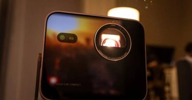 Vidéoprojecteur Leica Cine Compact 1 en boîtier aluminium noir, objectif Leica Summicron éclairé en rouge par la source triple laser RGB, posé sur un guéridon près d'une bougie dans une ambiance salon tamisée