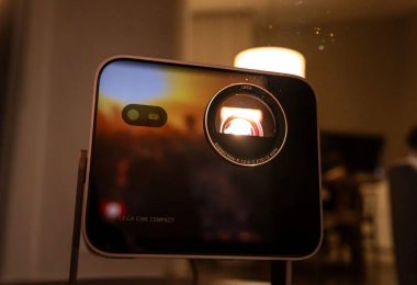 Vidéoprojecteur Leica Cine Compact 1 en boîtier aluminium noir, objectif Leica Summicron éclairé en rouge par la source triple laser RGB, posé sur un guéridon près d'une bougie dans une ambiance salon tamisée