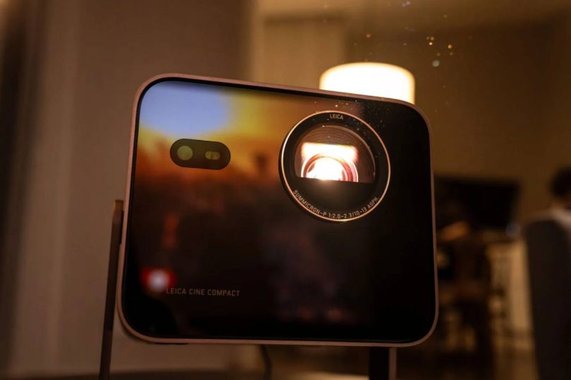 Vidéoprojecteur Leica Cine Compact 1 en boîtier aluminium noir, objectif Leica Summicron éclairé en rouge par la source triple laser RGB, posé sur un guéridon près d'une bougie dans une ambiance salon tamisée