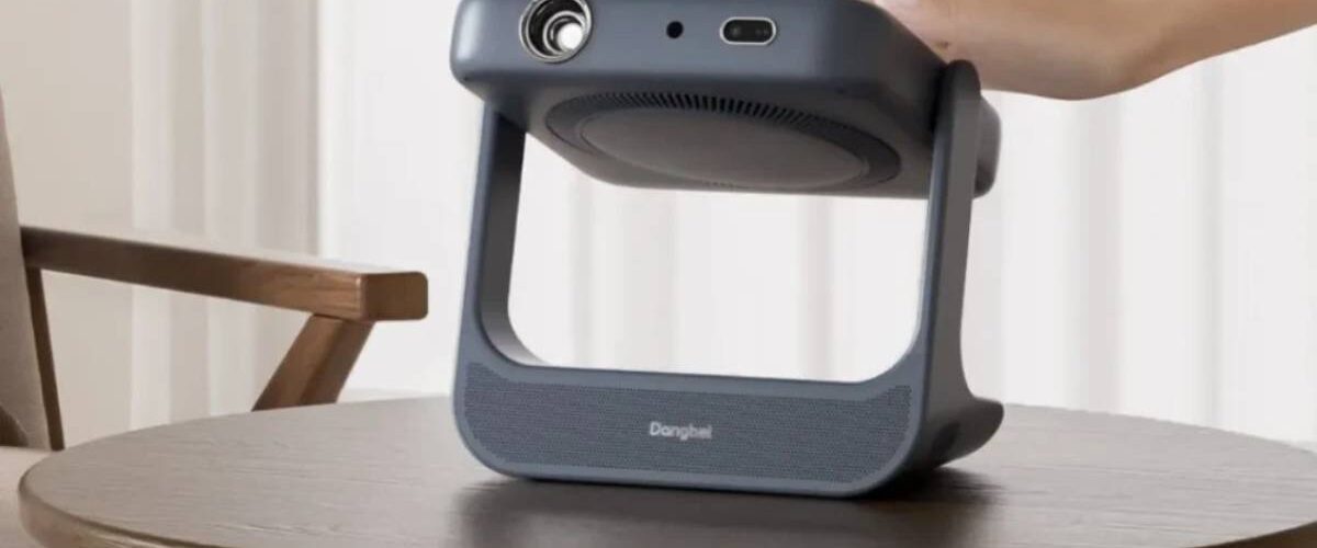 Vidéoprojecteur portable laser Dangbei RX10 gris anthracite posé sur une table basse en bois, bloc optique monté sur cardan pivotant dans un arceau poignée, main de l'utilisateur saisissant l'appareil dans un salon clair