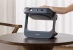Vidéoprojecteur portable laser Dangbei RX10 gris anthracite posé sur une table basse en bois, bloc optique monté sur cardan pivotant dans un arceau poignée, main de l'utilisateur saisissant l'appareil dans un salon clair