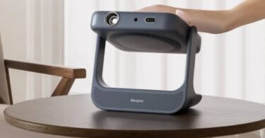Vidéoprojecteur portable laser Dangbei RX10 gris anthracite posé sur une table basse en bois, bloc optique monté sur cardan pivotant dans un arceau poignée, main de l'utilisateur saisissant l'appareil dans un salon clair