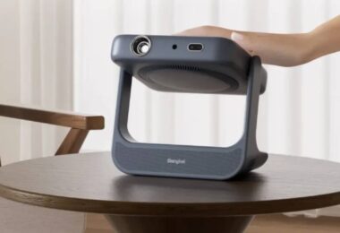 Vidéoprojecteur portable laser Dangbei RX10 gris anthracite posé sur une table basse en bois, bloc optique monté sur cardan pivotant dans un arceau poignée, main de l'utilisateur saisissant l'appareil dans un salon clair
