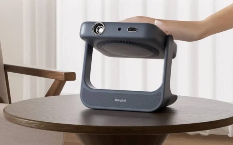 Vidéoprojecteur portable laser Dangbei RX10 gris anthracite posé sur une table basse en bois, bloc optique monté sur cardan pivotant dans un arceau poignée, main de l'utilisateur saisissant l'appareil dans un salon clair