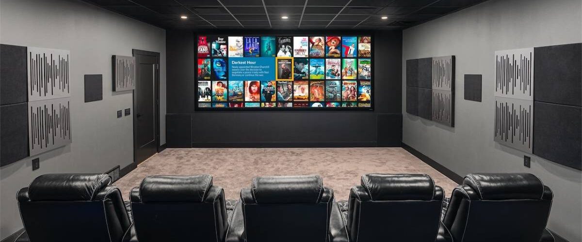 Salle de home cinéma privée traitée acoustiquement, grand écran de projection large affichant une interface de bibliothèque de films, deux rangées de fauteuils inclinables en cuir noir, panneaux absorbants et diffuseurs muraux, plafond noir avec spots encastrés