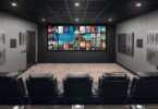 Salle de home cinéma privée traitée acoustiquement, grand écran de projection large affichant une interface de bibliothèque de films, deux rangées de fauteuils inclinables en cuir noir, panneaux absorbants et diffuseurs muraux, plafond noir avec spots encastrés