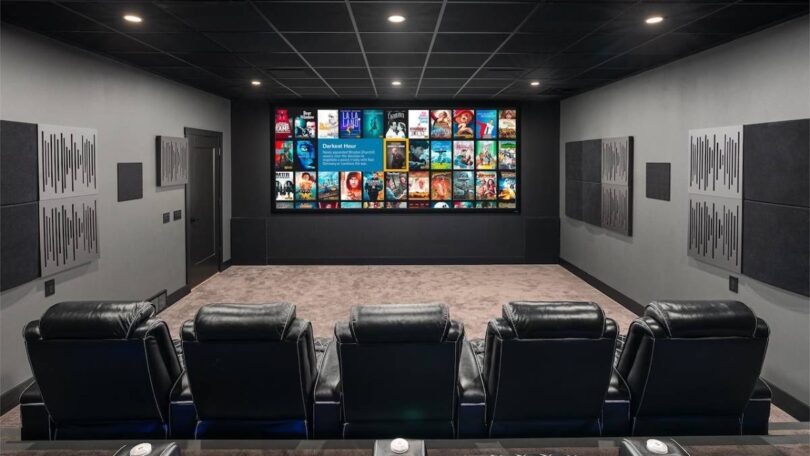 Salle de home cinéma privée traitée acoustiquement, grand écran de projection large affichant une interface de bibliothèque de films, deux rangées de fauteuils inclinables en cuir noir, panneaux absorbants et diffuseurs muraux, plafond noir avec spots encastrés