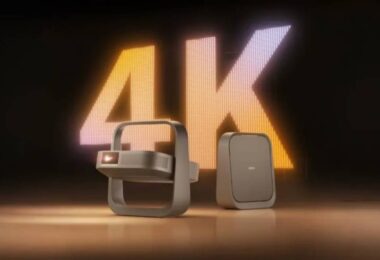 Projecteur portable XGIMI Z9X 4K High Brightness Edition au design cadre-poignée cardan, finition bronze, posé à côté de son bloc d'alimentation devant un mur LED géant affichant 4K en lettres lumineuses orange et blanches