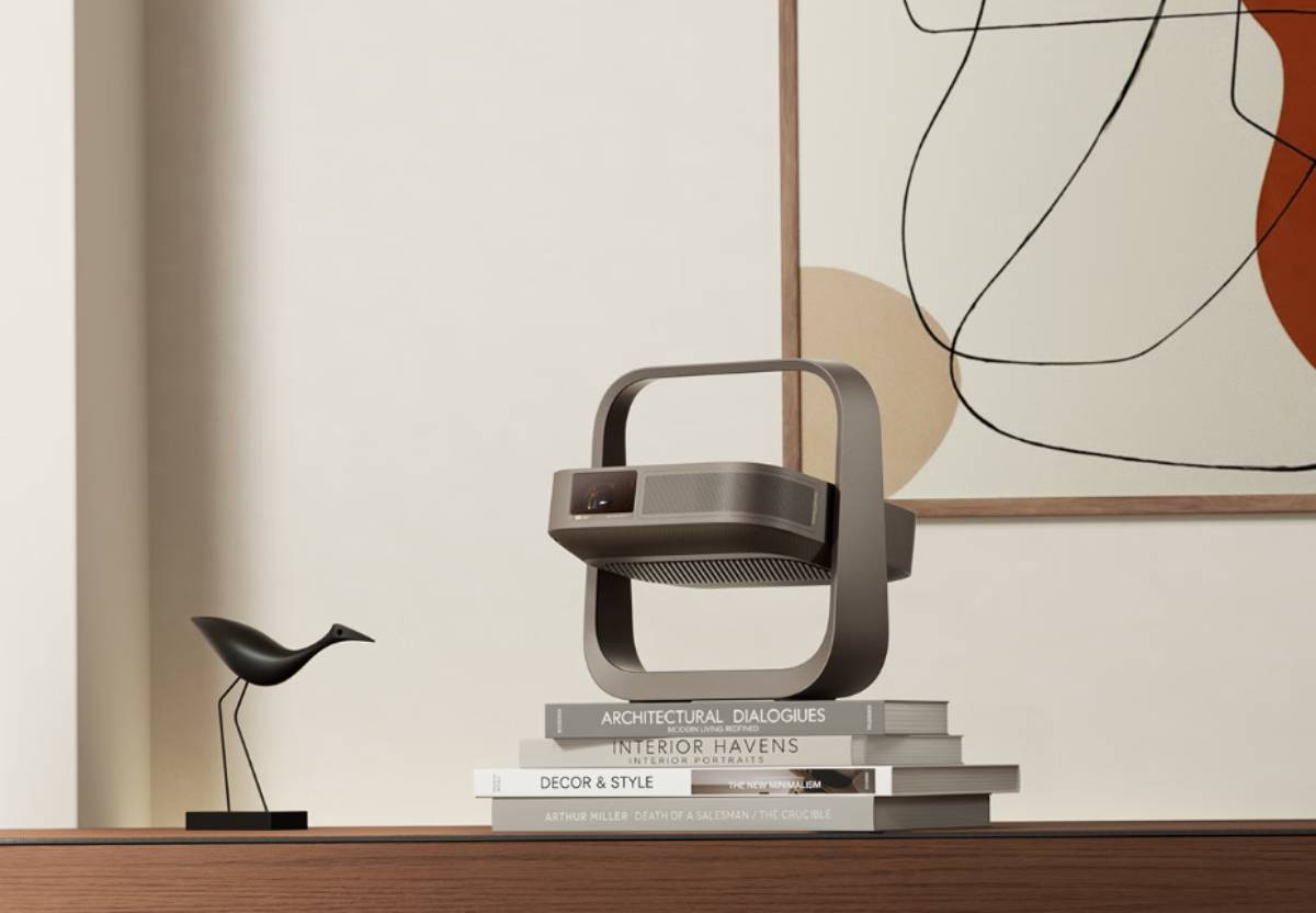 Projecteur portable XGIMI Z9X 4K finition bronze taupe posé sur une pile de livres de design dans un salon minimaliste, cadre-poignée cardan incliné vers le haut, enfilade en bois clair, sculpture d'oiseau noire et tableau abstrait au mur
