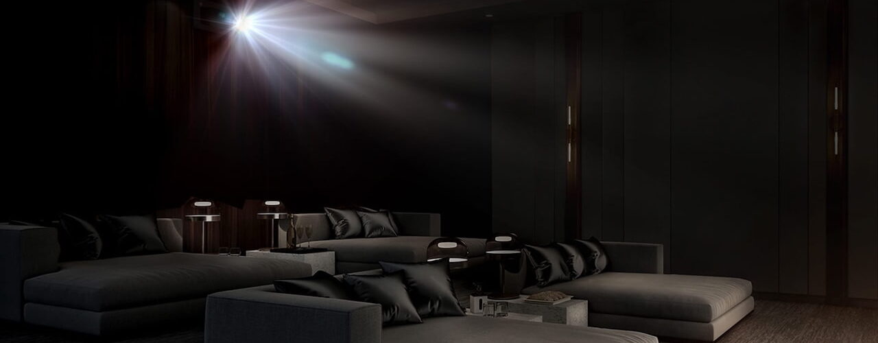 Faisceau lumineux d'un vidéoprojecteur laser 4K type Epson QL7000 traversant une salle de cinéma privée sombre équipée de larges banquettes grises, illustrant la projection haute luminosité de 10 000 lumens sur très grand écran.