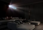 Faisceau lumineux d'un vidéoprojecteur laser 4K type Epson QL7000 traversant une salle de cinéma privée sombre équipée de larges banquettes grises, illustrant la projection haute luminosité de 10 000 lumens sur très grand écran.