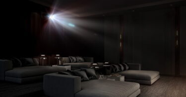 Faisceau lumineux d'un vidéoprojecteur laser 4K type Epson QL7000 traversant une salle de cinéma privée sombre équipée de larges banquettes grises, illustrant la projection haute luminosité de 10 000 lumens sur très grand écran.