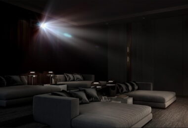 Faisceau lumineux d'un vidéoprojecteur laser 4K type Epson QL7000 traversant une salle de cinéma privée sombre équipée de larges banquettes grises, illustrant la projection haute luminosité de 10 000 lumens sur très grand écran.