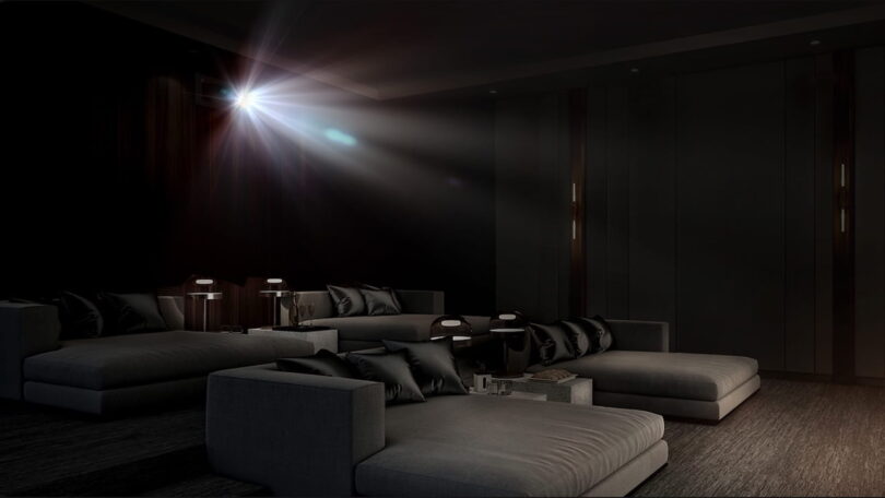 Faisceau lumineux d'un vidéoprojecteur laser 4K type Epson QL7000 traversant une salle de cinéma privée sombre équipée de larges banquettes grises, illustrant la projection haute luminosité de 10 000 lumens sur très grand écran.