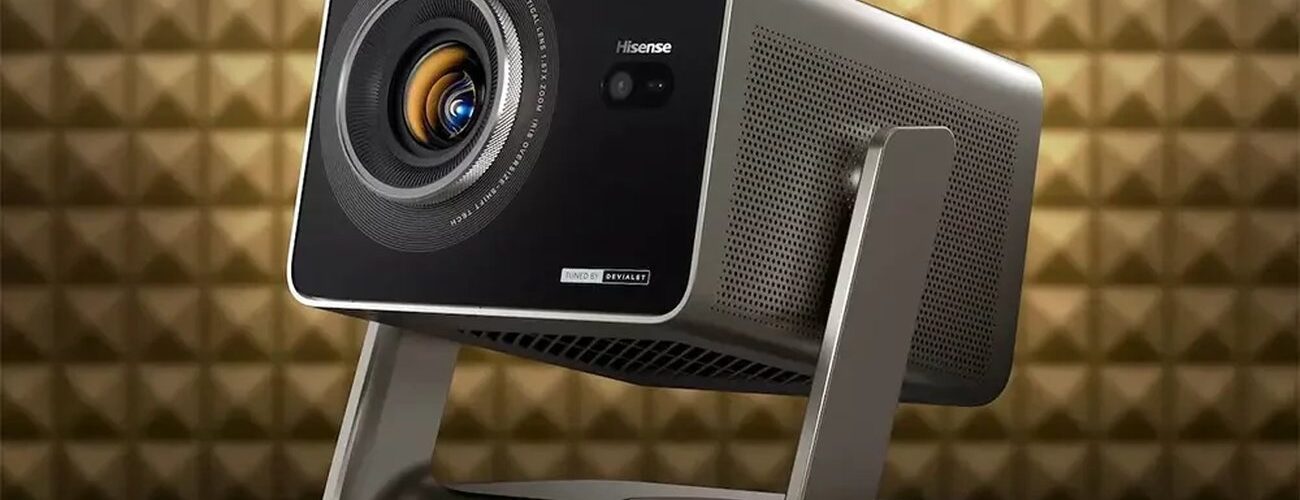 Projecteur Hisense de la gamme C3 triple laser 4K, finition gris titane sur support à cardan, objectif zoom 1,6x avec iris et marquage Tuned by Devialet, devant un mur acoustique doré