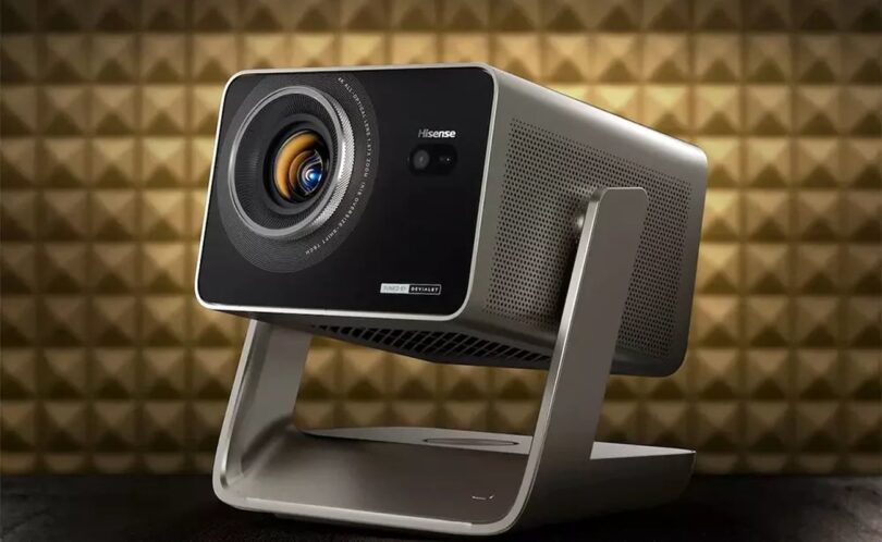 Projecteur Hisense de la gamme C3 triple laser 4K, finition gris titane sur support à cardan, objectif zoom 1,6x avec iris et marquage Tuned by Devialet, devant un mur acoustique doré