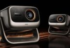 Deux vidéoprojecteurs JMGO Iris triple laser 4K 120 Hz, coque argent champagne, façade noire, objectif cerclé d'orange et rotule en U, sur fond noir avec étincelles cuivrées