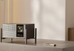 Vidéoprojecteur 4K XGIMI TITAN Noir posé sur son pied à quatre colonnes sur un meuble en bois gris, façade grille aluminium, objectif zoom, logos Harman Kardon et IMAX Enhanced, télécommande métal