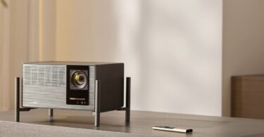 Vidéoprojecteur 4K XGIMI TITAN Noir posé sur son pied à quatre colonnes sur un meuble en bois gris, façade grille aluminium, objectif zoom, logos Harman Kardon et IMAX Enhanced, télécommande métal