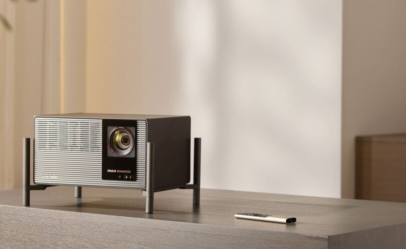 Vidéoprojecteur 4K XGIMI TITAN Noir posé sur son pied à quatre colonnes sur un meuble en bois gris, façade grille aluminium, objectif zoom, logos Harman Kardon et IMAX Enhanced, télécommande métal